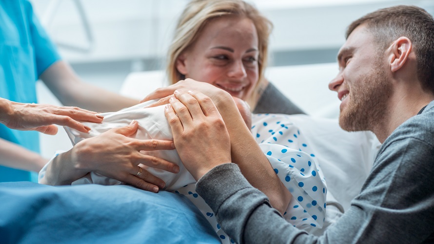 Junges Paar nach Entbindung Junge Frau haelt glücklich Neugeborenes nach Geburt auf ihrem Bauch, Partner legt Hand auf Baby und lächelt
